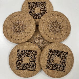 May include: Set of five cork coasters with a black and white mandala design and the text "alchemist organics" on two of the coasters. The other three coasters have a black and white QR code.