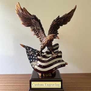 May include: A bronze eagle sculpture with outstretched wings holding an American flag. The eagle is perched on a wooden base with a gold plaque that reads "Custom Engraving".