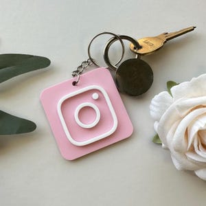 May include: Pink Instagram logo keychain with a white outline. The keychain is attached to a silver key ring with a key and a silver circle.
