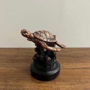 May include: A small copper-colored turtle figurine on a black base. The turtle is swimming above a coral reef.