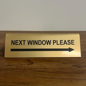 May include: Gold-colored desk sign with black text that reads "NEXT WINDOW PLEASE" and a black arrow pointing to the right.