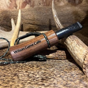 May include: A wooden Wilson Game Calls duck call with a black mouthpiece and a green and black lanyard. The call is made of dark brown wood and is resting on a textured surface with antlers in the background.