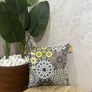 Könnte beinhalten: Ein quadratisches Zierkissen mit einem Patchwork-Design aus geometrischen und floralen Mustern in Gelb-, Grau-, Schwarz- und Weißtönen. Das Kissen ist an den beiden oberen Ecken mit kleinen goldfarbenen Quasten versehen.