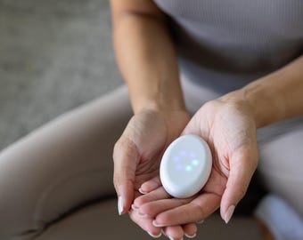 May include: A smooth, white, oval-shaped handheld device featuring a cluster of small, glowing circular lights on its surface. The device has a minimalist, rounded form and is designed to fit comfortably within the palm of a hand for tactile use.
