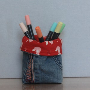 May include: A denim pencil holder with a red fabric lining featuring a pink animal print. The holder is filled with colourful markers.