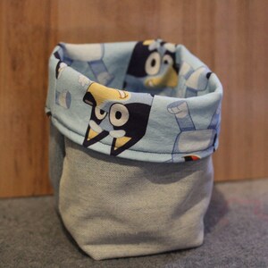 May include: A blue and white fabric storage bin with a cartoon character design. The bin is made of denim and has a rolled top.