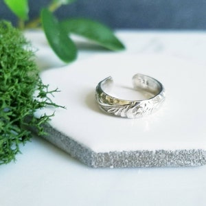 May include: A silver toe ring with a floral pattern. The ring is open at the top and has a textured surface. The ring is displayed on a white hexagonal tile.