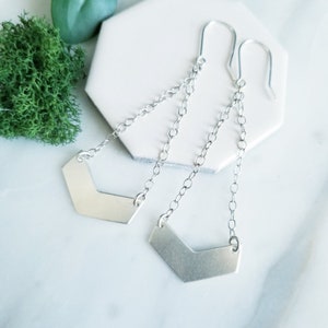 May include: A pair of silver earrings with a chevron-shaped pendant hanging from a delicate chain. The earrings are made of sterling silver and have a simple, modern design.