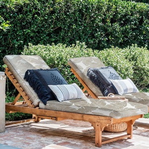 May include: Two wooden lounge chairs with beige cushions and leaf patterns. The chairs are positioned on a brick patio with a green hedge in the background. Each chair has two pillows, one black with fringe and one white with black stripes.