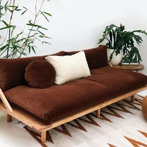 May include: A contemporary sofa with a wooden frame and a deep brown, plush cushion. It has a round and rectangular pillow, and a white pillow. The sofa is in a room with plants and a patterned rug.