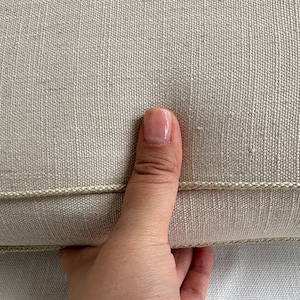 Puede incluir: Primer plano de un coj&iacute;n de lino beige con un ribete decorativo de cord&oacute;n. El coj&iacute;n tiene una superficie texturizada y una paleta de colores neutros, adecuada para varios estilos de decoraci&oacute;n del hogar. Una mano sostiene el coj&iacute;n.
