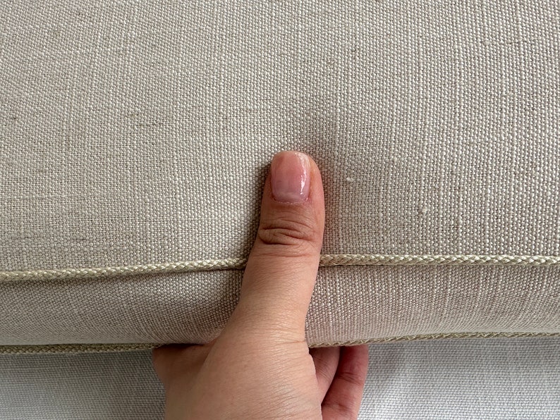 Puede incluir: Primer plano de un coj&iacute;n de lino beige con un ribete decorativo de cord&oacute;n. El coj&iacute;n tiene una superficie texturizada y una paleta de colores neutros, adecuada para varios estilos de decoraci&oacute;n del hogar. Una mano sostiene el coj&iacute;n.