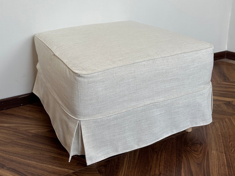 Op de afbeelding: Een witte linnen ottomaan met een geplooide rok. De ottomaan staat op een houten vloer.