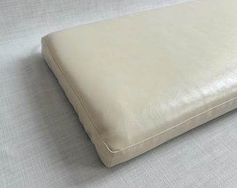 Pode incluir: Uma almofada retangular com uma superfície lisa de cor branco-sujo, semelhante a couro. A almofada apresenta costuras retas e limpas ao longo das bordas, criando uma forma estruturada e quadrada com um brilho subtil no painel superior.
