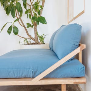 Op de afbeelding: Een lichtblauwe daybed in futonstijl met een natuurhouten frame. De daybed heeft twee grote, bijpassende kussens en een aangepaste matras. Een potplant met groene bladeren staat op de achtergrond.