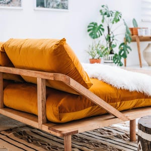 May include: A contemporary daybed featuring a wooden frame and mustard-yellow cushions. A fluffy white throw blanket is draped over the cushions. This daybed is a stylish piece of furniture for a living room or bedroom.