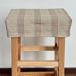 Puede incluir: Un taburete de madera con una funda de asiento de tela a cuadros marrón y beige. El taburete tiene tres estantes.