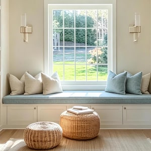 Può includere: Una comoda seduta vicino alla finestra con un cuscino azzurro e diversi cuscini. Due pouf intrecciati sono sul pavimento in legno. La finestra è incorniciata da finiture bianche e si affaccia su un prato verde.