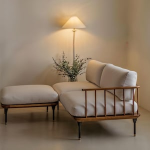 Puede incluir: Un moderno juego de sofá y otomana de color crema con patas de madera y metal negro. El sofá tiene un reposabrazos de madera con husillos verticales. Una lámpara de pie con una pantalla beige y un jarrón de vegetación están en el fondo.