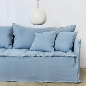 May include: A light blue sofa with matching throw pillows. The sofa has a slipcover with white piping along the edges. A white, spherical light fixture hangs above the sofa.