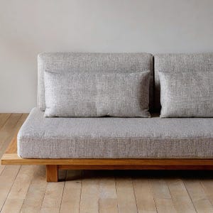 Op de afbeelding: Een moderne grijze bank met twee rugkussens en een rechthoekig lendenkussen. De bank heeft een houten basis en staat op een houten vloer. De kussens en de bank zijn bedekt met een grijze stof met textuur.