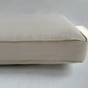 Puede incluir: Primer plano de un coj&iacute;n rectangular beige con una tela clara y texturizada. Los bordes est&aacute;n cuidadosamente acabados con un ribete fino a juego. El coj&iacute;n parece nuevo y sin usar.