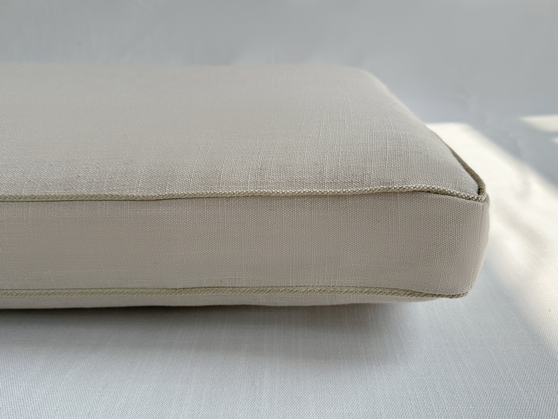 Puede incluir: Primer plano de un coj&iacute;n rectangular beige con una tela clara y texturizada. Los bordes est&aacute;n cuidadosamente acabados con un ribete fino a juego. El coj&iacute;n parece nuevo y sin usar.