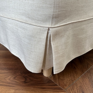 Op de afbeelding: Een close-up van de onderkant van een beige linnen stoelhoes met een geplooid detail. De hoes zit op een houten stoelpoot en de stoel staat op een donkere houten vloer.