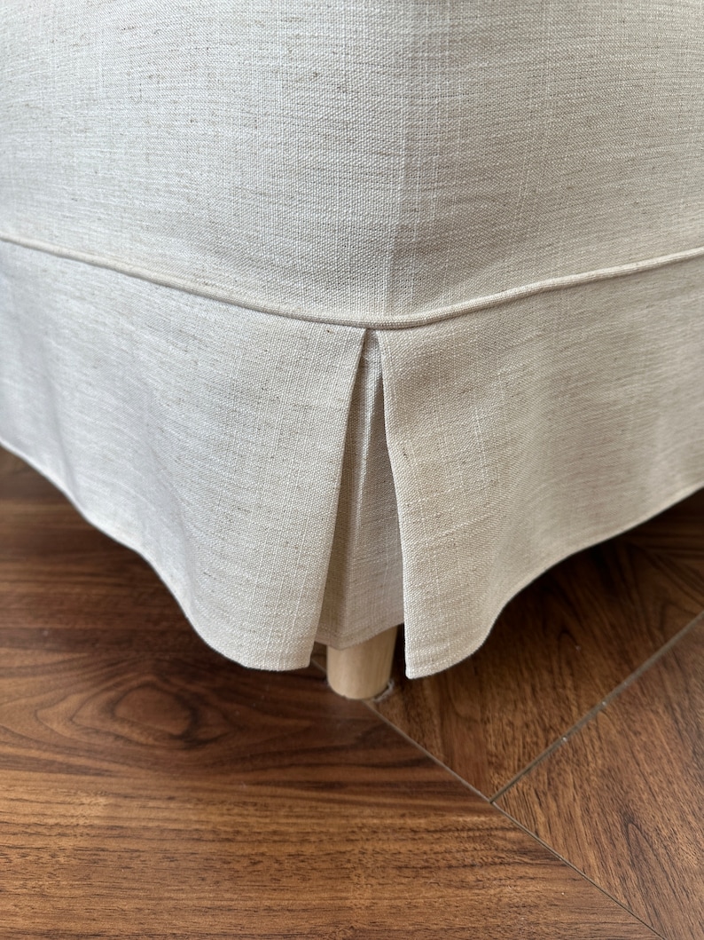 Op de afbeelding: Een close-up van de onderkant van een beige linnen stoelhoes met een geplooid detail. De hoes zit op een houten stoelpoot en de stoel staat op een donkere houten vloer.