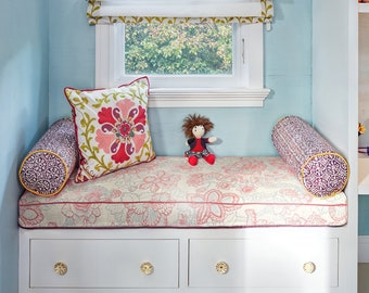 May include: A white window seat with a floral patterned cushion and two purple and gold patterned cylindrical pillows. A doll is sitting on the cushion.