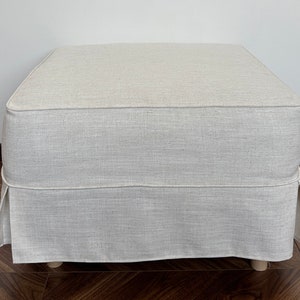 Op de afbeelding: Een vierkante, cr&egrave;mekleurige ottomaan met een bekleding van linnen stof en een geplooide rok. De ottomaan staat op een houten vloer.