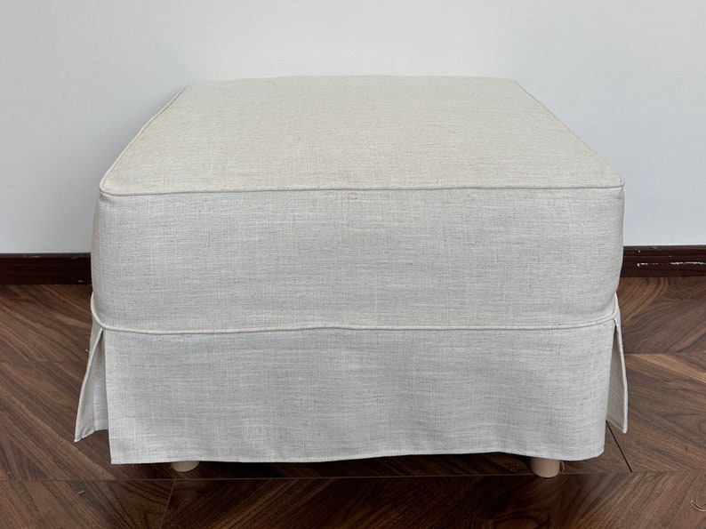 Op de afbeelding: Een vierkante, cr&egrave;mekleurige ottomaan met een bekleding van linnen stof en een geplooide rok. De ottomaan staat op een houten vloer.