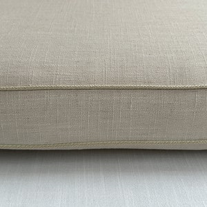 Puede incluir: Primer plano de un coj&iacute;n de lino beige con una superficie texturizada. El coj&iacute;n tiene un borde cosido de color claro. La tela parece estar tejida apretadamente, lo que sugiere durabilidad. El color neutro lo hace adecuado para varios estilos de decoraci&oacute;n.