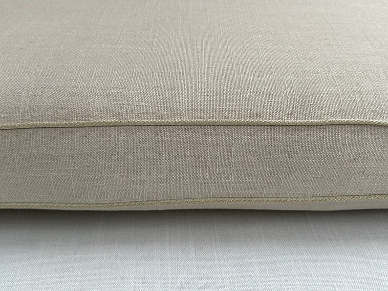 Puede incluir: Primer plano de un coj&iacute;n de lino beige con una superficie texturizada. El coj&iacute;n tiene un borde cosido de color claro. La tela parece estar tejida apretadamente, lo que sugiere durabilidad. El color neutro lo hace adecuado para varios estilos de decoraci&oacute;n.