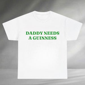 A white t-shirt with the text "DADDA NEEDS A GUINNESS" printed on it in green.