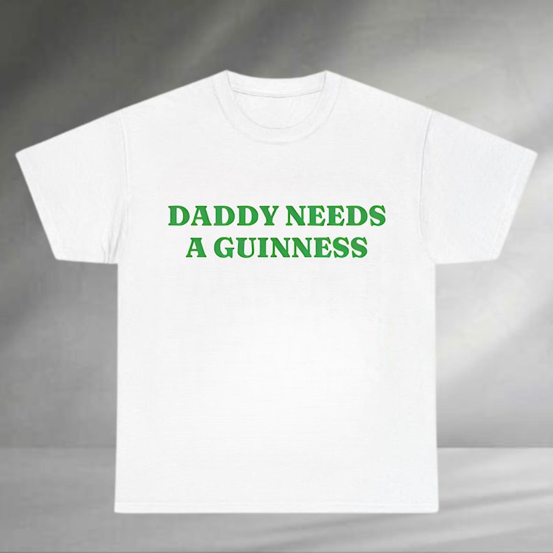 A white t-shirt with the text "DADDA NEEDS A GUINNESS" printed on it in green.