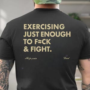 May include: Black t-shirt with the text "EXERCISING JUST ENOUGH TO F**K & FIGHT." in a gold color. Below the text are the words "Not for pussies" and "Frazed". The shirt is a crew neck.