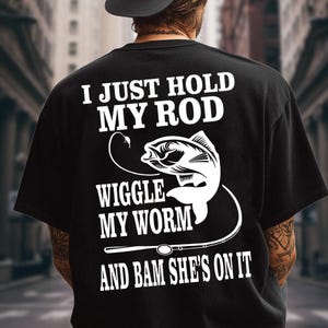 May include: A black t-shirt featuring white screen-printed text on the back that reads I JUST HOLD MY ROD WIGGLE MY WORM AND BAM SHE'S ON IT, accompanied by a graphic illustration of a fish hooked on a fishing line and a fishing rod.