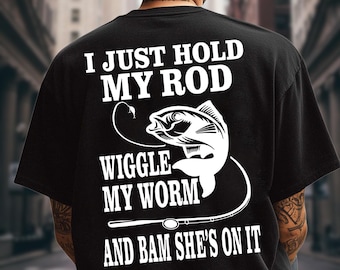 May include: A black t-shirt featuring white screen-printed text on the back that reads I JUST HOLD MY ROD WIGGLE MY WORM AND BAM SHE'S ON IT, accompanied by a graphic illustration of a fish hooked on a fishing line and a fishing rod.