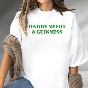 A person wearing a white t-shirt with the text "DADDY NEEDS A GUINNESS" printed on it.