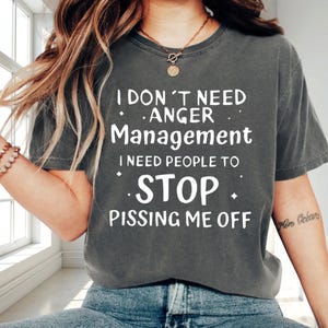 May include: Dark gray t-shirt with the text "I DON'T NEED ANGER Management I NEED PEOPLE TO STOP PISSING ME OFF" in white. The shirt is a casual style, suitable for everyday wear.