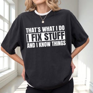 May include: Black t-shirt with the text "THAT'S WHAT I DO I FIX STUFF AND I KNOW THINGS" in white block letters. The shirt has a white crew neck collar and is worn by a person.