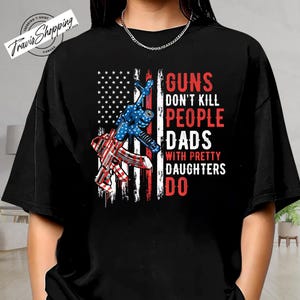 May include: Black t-shirt featuring a graphic design with an American flag, two guns, and the text "GUNS DON'T KILL PEOPLE DADS WITH PRETTY DAUGHTERS DO." The shirt also has the text "TRENDING T-SHIRT" and "Travis Shopping CANADA."