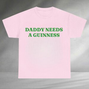A pink t-shirt with the text "DADDAY NEEDS A GUINNESS" printed on it in green letters.