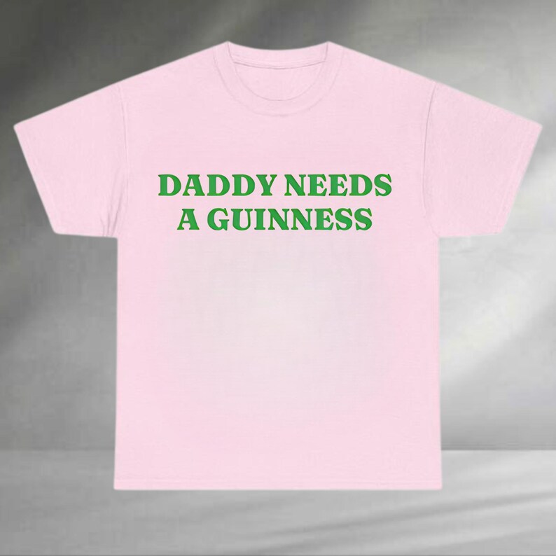 A pink t-shirt with the text "DADDAY NEEDS A GUINNESS" printed on it in green letters.