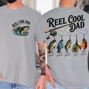 A gray t-shirt with a graphic design on the front and back, featuring a fishing rod and fish illustrations. The design includes the text "REEL COOL DAD" and names of people associated with the design.