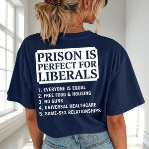 May include: A navy blue short-sleeved t-shirt featuring white text on the back that reads: PRISON IS PERFECT FOR LIBERALS, followed by a numbered list: 1. EVERYONE IS EQUAL, 2. FREE FOOD & HOUSING, 3. NO GUNS, 4. UNIVERSAL HEALTHCARE, 5. SAME-SEX RELATIONSHIPS.