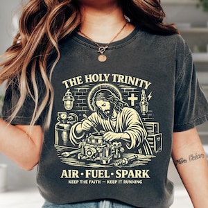 May include: Dark gray t-shirt featuring a graphic of Jesus working on an engine carburetor, with the text "THE HOLY TRINITY" above and "AIR FUEL SPARK" below. The shirt also includes the phrases "KEEP THE FAITH - KEEP IT RUNNING".