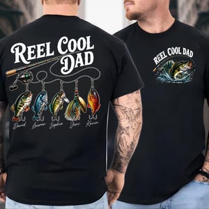 a black t-shirt with a design that includes a fishing rod, fish, and the text "Reel Cool Dad" printed on it.