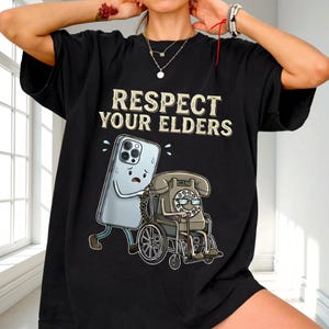 A person wearing a black t-shirt with a graphic design depicting a phone and a wheelchair, along with the text "RESPECT YOUR ELDERS".